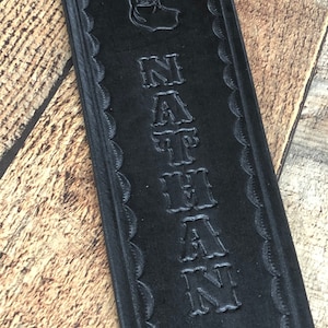 May include: Black leather strap with a deer head design and the name "NATHAN" embossed in a decorative font.