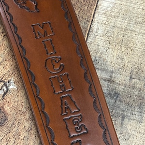 May include: Brown leather strap with a decorative border and the name "MICHAEL" embossed in a vintage font. A deer head is also embossed at the top of the strap.
