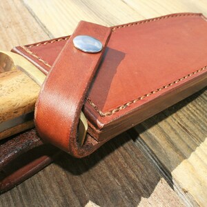 May include: Close-up of a brown leather knife sheath with a wooden handle knife partially inserted. The sheath has a strap with a silver button closure. The knife handle is light brown with a wood grain pattern. The sheath is stitched with light brown thread.