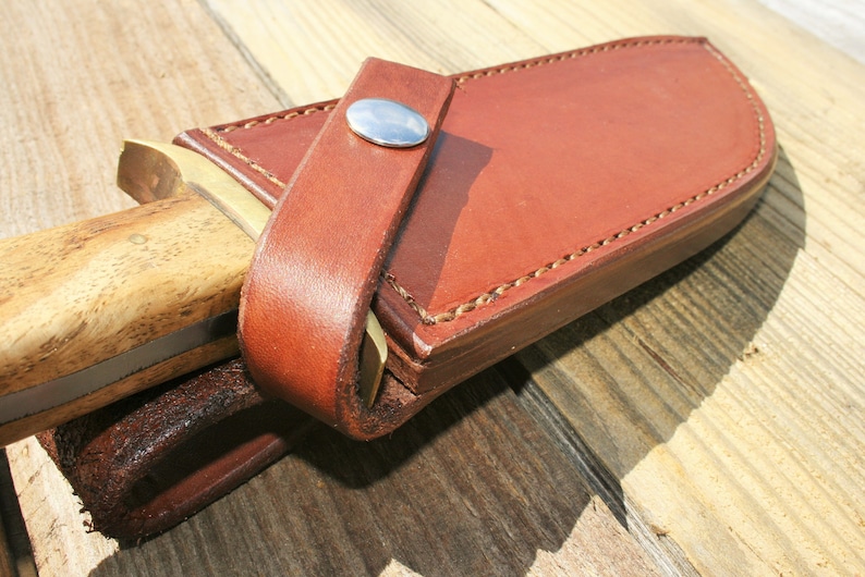 May include: Close-up of a brown leather knife sheath with a wooden handle knife partially inserted. The sheath has a strap with a silver button closure. The knife handle is light brown with a wood grain pattern. The sheath is stitched with light brown thread.
