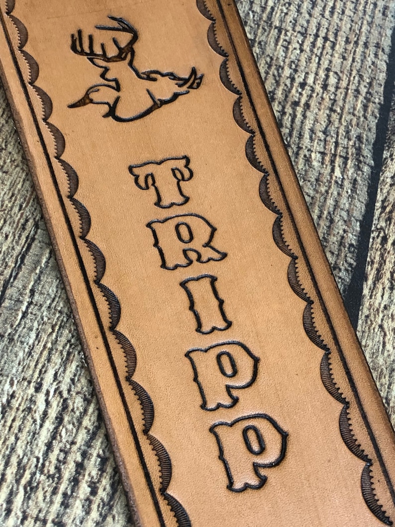 May include: A brown leather strap with a decorative border and the name "TRIPP" embossed in a stylized font. A silhouette of a flying duck with antlers is also embossed on the strap.