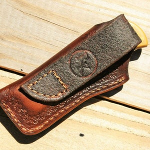 May include: A brown leather knife sheath with a stitched flap and a brown leather belt loop. The sheath has a decorative stitching pattern and a small, circular logo.