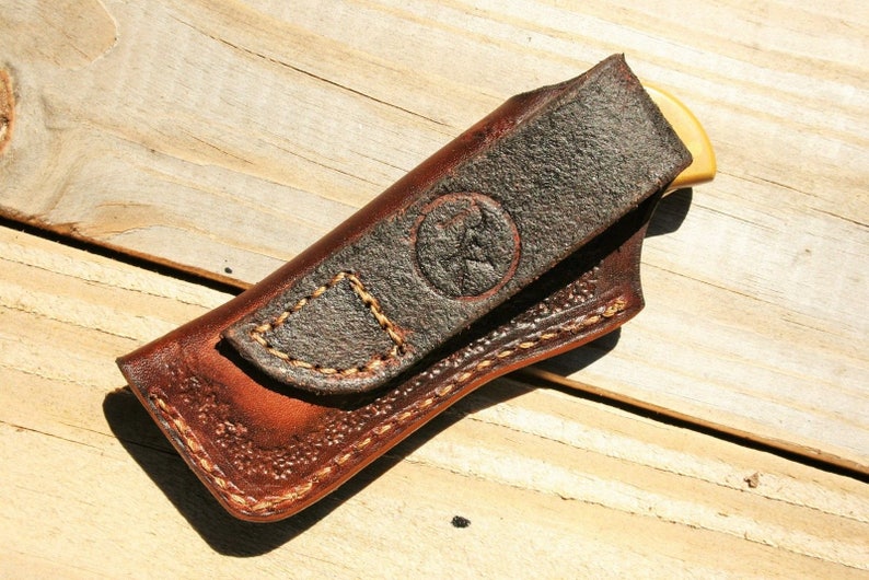 May include: A brown leather knife sheath with a stitched flap and a brown leather belt loop. The sheath has a decorative stitching pattern and a small, circular logo.