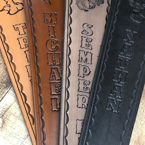 May include: Four leather straps with different designs and names. The first strap is light brown with a deer design and the name "TRILL" engraved. The second strap is brown with a deer design and the name "MICHAEL" engraved. The third strap is gray with a military emblem and the name "SEMPER FI" engraved. The fourth strap is black with a deer design and the name "NATHAN" engraved.