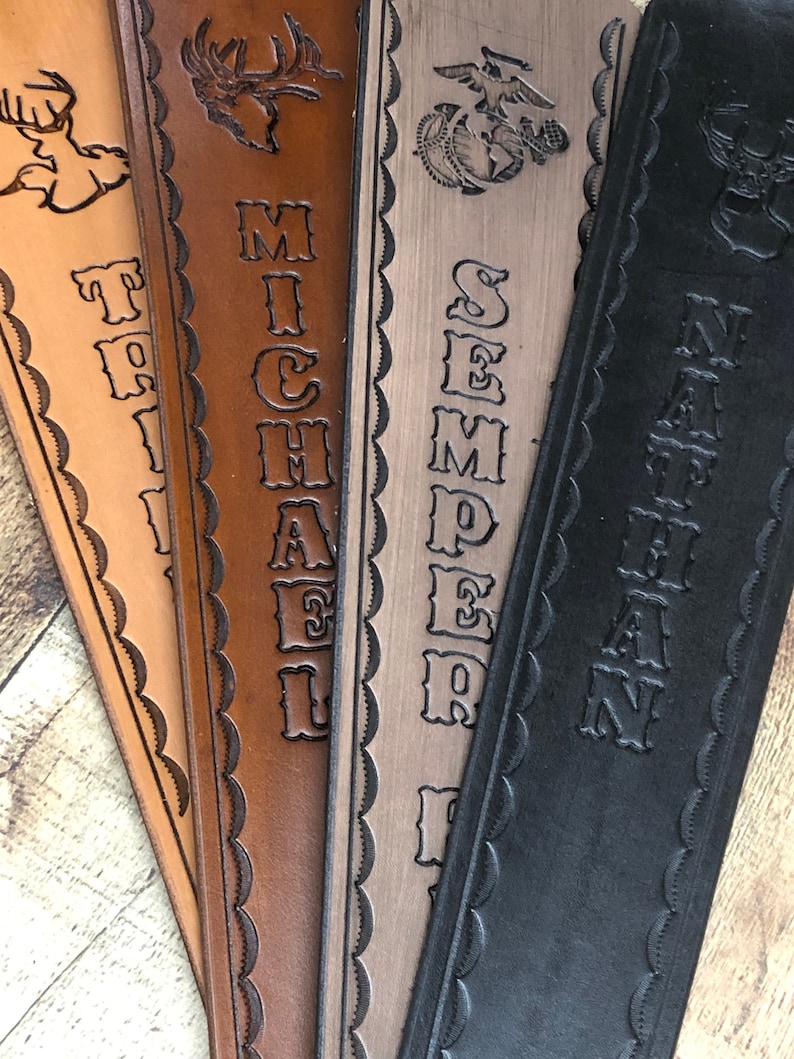May include: Four leather straps with different designs and names. The first strap is light brown with a deer design and the name "TRILL" engraved. The second strap is brown with a deer design and the name "MICHAEL" engraved. The third strap is gray with a military emblem and the name "SEMPER FI" engraved. The fourth strap is black with a deer design and the name "NATHAN" engraved.