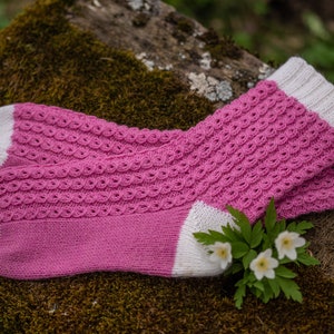 May include: A pair of pink and white knitted socks with a textured pattern. The socks are lying on a bed of moss.