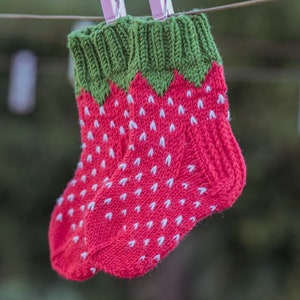 Puede incluir: Un par de calcetines tejidos con un diseño de fresa. Los calcetines son rojos con semillas blancas y hojas verdes. Están colgados en un tendedero con pinzas de ropa rosas.