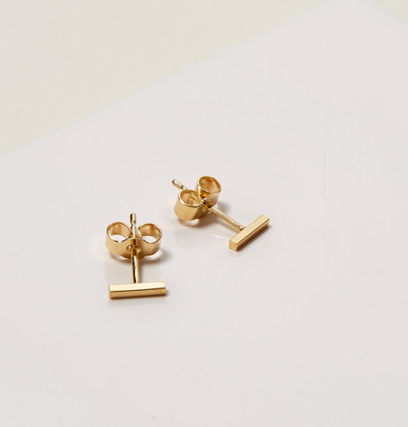 May include: A pair of gold bar stud earrings. The earrings are simple and minimalist, with a small, rectangular bar.