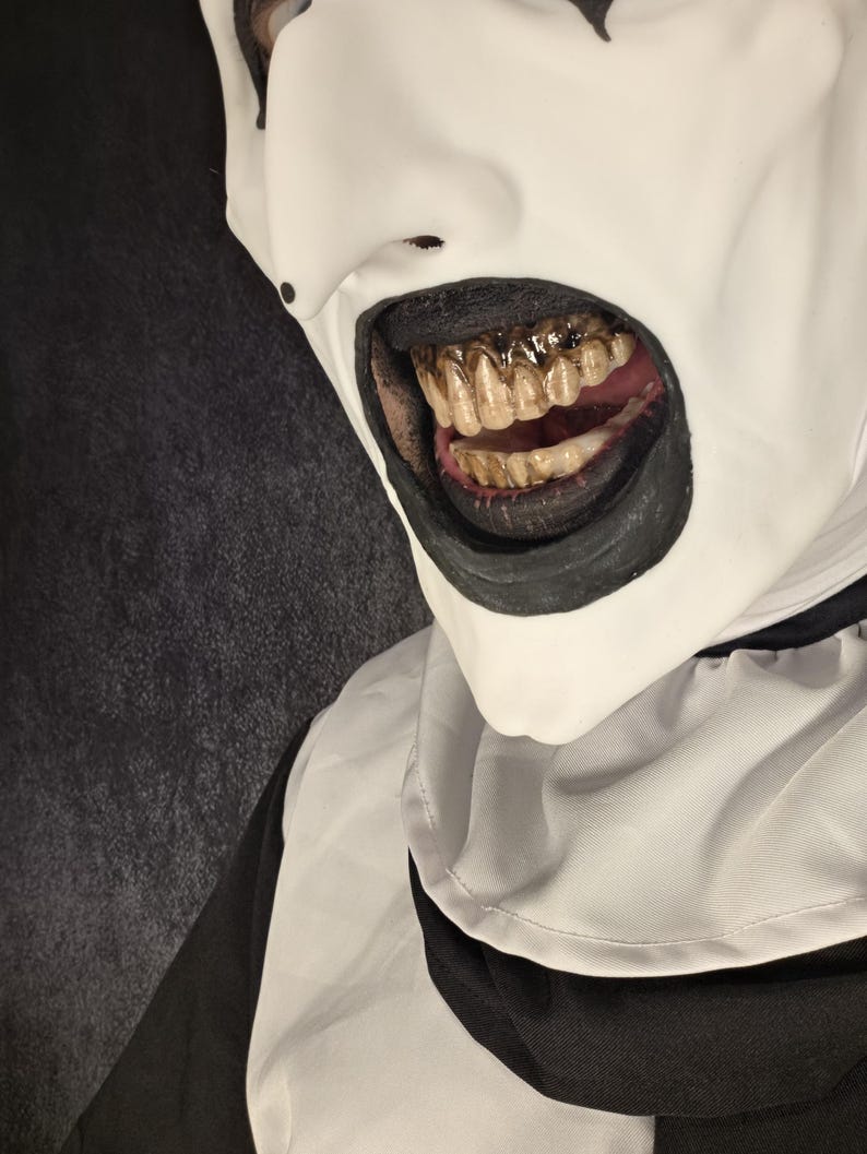 May include: A full-head mask featuring a smooth white face with black markings around the eyes and mouth. The mouth is open, revealing a set of stained, yellowed teeth. The mask includes a white collar and black fabric sections at the neck.