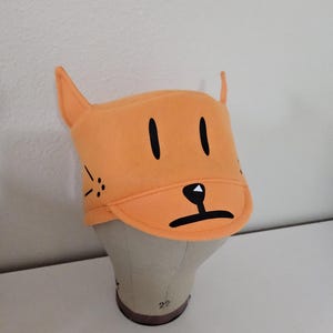 May include: Orange cat-themed hat with black facial features, including eyes, nose, and whiskers. The hat has upright ears and a small brim. The hat is made of a soft material and is designed to resemble a cartoon cat.