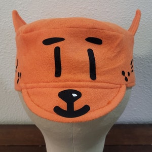 May include: Orange felt hat with black cartoon face and ears. The hat has a brim and a simple design.