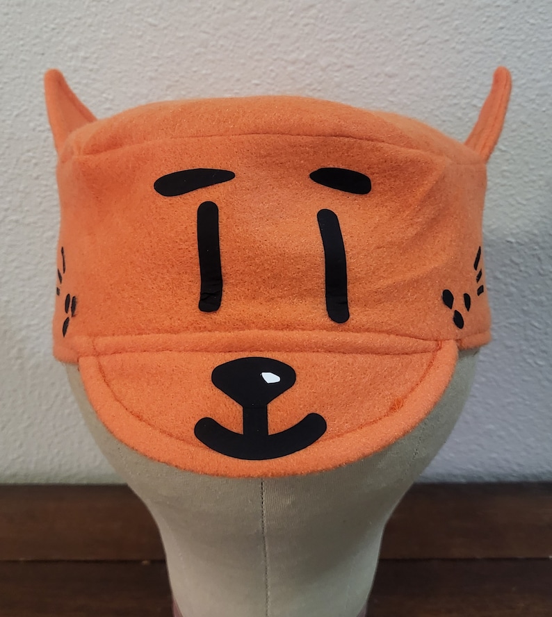 May include: Orange felt hat with black cartoon face and ears. The hat has a brim and a simple design.