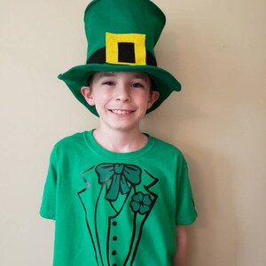 May include: A young person wearing a green t-shirt with a black tuxedo design and a green leprechaun hat with a yellow buckle.