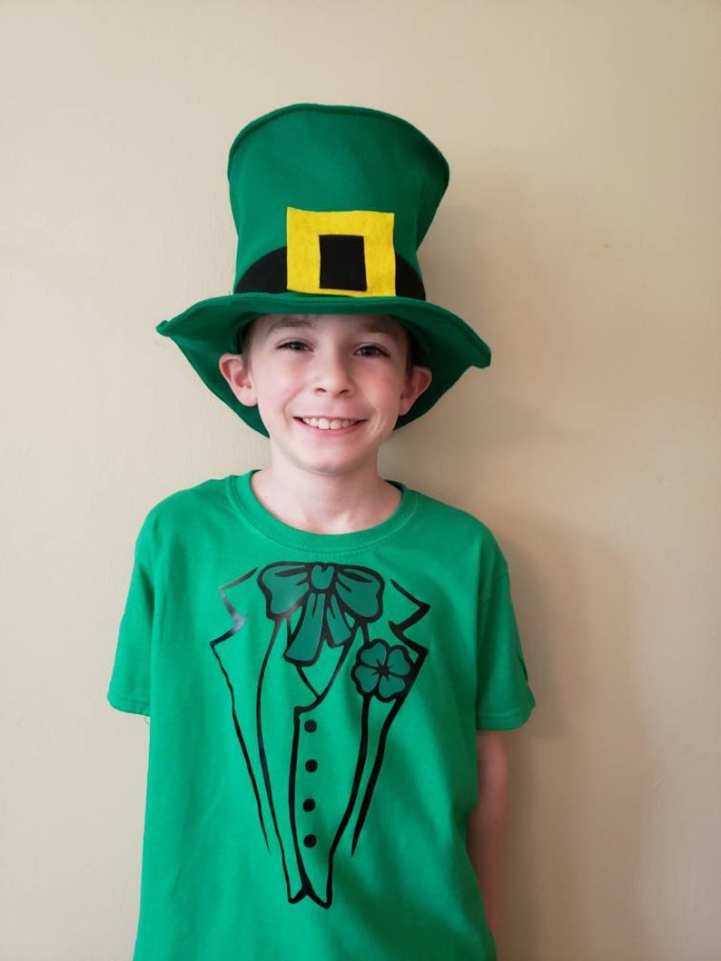 May include: A young person wearing a green t-shirt with a black tuxedo design and a green leprechaun hat with a yellow buckle.