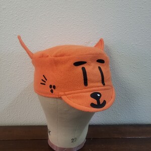 May include: Orange felt hat with a black cat face design. The hat has a brim and two cat ears.