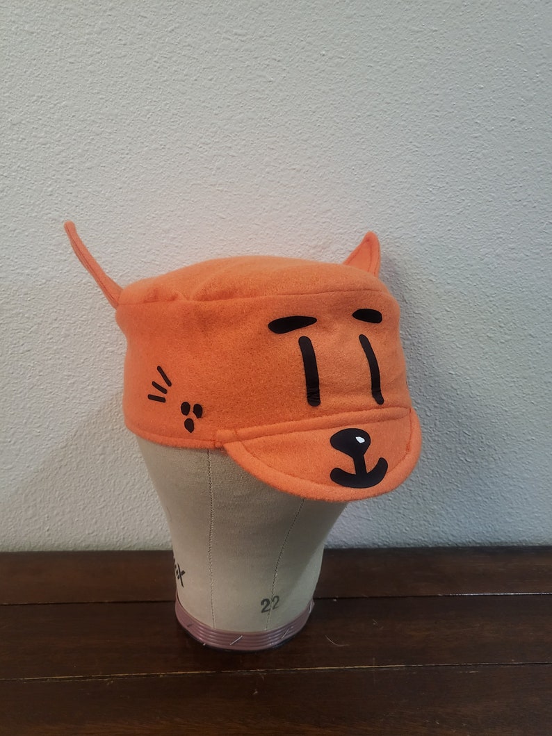May include: Orange felt hat with a black cat face design. The hat has a brim and two cat ears.
