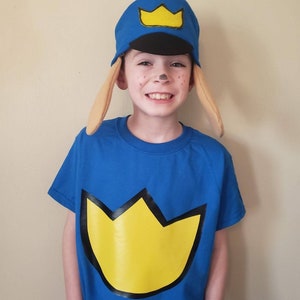 May include: A child wearing a blue t-shirt with a yellow crown design and a blue and yellow hat with a crown design. The child is also wearing a pair of brown felt ears.