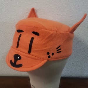 May include: Orange felt hat with black cat ears and a black cat face with eyes, nose, and whiskers.