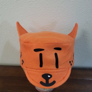 May include: Orange fleece hat with black felt appliques of a cat face. The hat has two pointy ears and a brim.