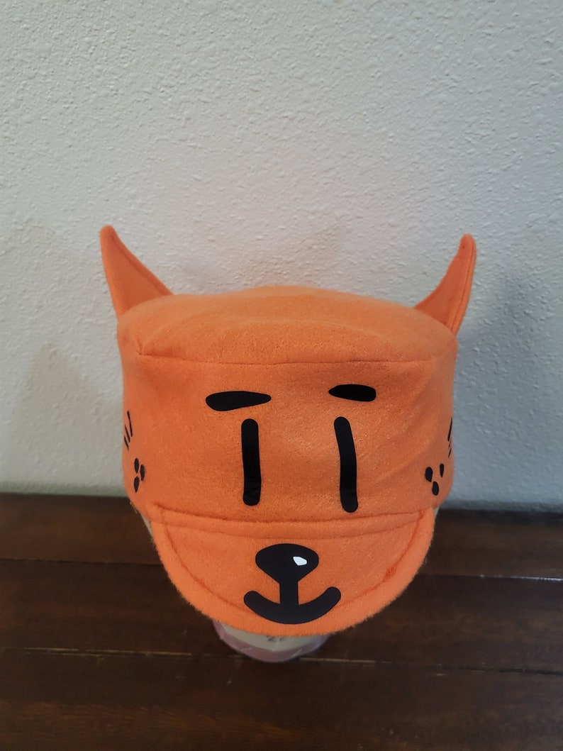 May include: Orange fleece hat with black felt appliques of a cat face. The hat has two pointy ears and a brim.