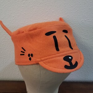 May include: Orange felt hat with black cat face design. The hat has a small brim and a pointed ear on the side.