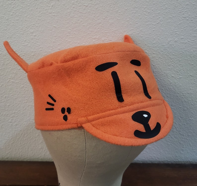 May include: Orange felt hat with black cat face design. The hat has a small brim and a pointed ear on the side.