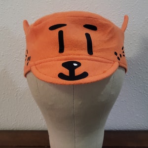 May include: Orange fleece hat with a black cartoon face on the front. The face has two eyes, a nose, and a mouth.