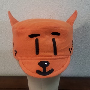 May include: Orange fleece hat with black felt appliques of a cat face. The hat has two pointy ears on top and a brim.