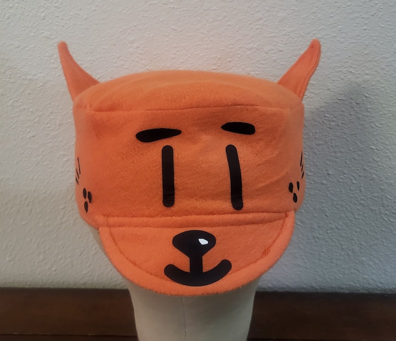 May include: Orange fleece hat with black felt appliques of a cat face. The hat has two pointy ears on top and a brim.