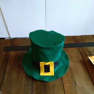 May include: A green felt leprechaun hat with a black band and a yellow buckle. The hat is sitting on a wooden surface.