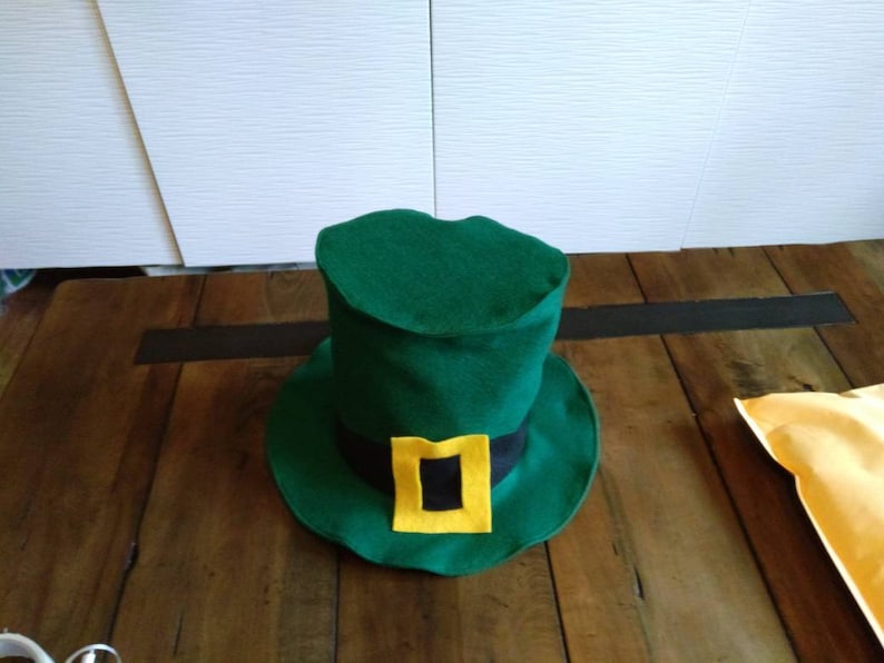 May include: A green felt leprechaun hat with a black band and a yellow buckle. The hat is sitting on a wooden surface.