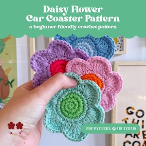 May include: A collection of handmade crochet daisy flower car coasters in various colors, including blue, purple, pink, and green. The coasters have a textured appearance and are held in a hand. The image includes text that reads "Daisy Flower Car Coaster Pattern".