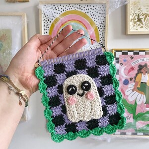 May include: Crocheted wall hanging featuring a friendly ghost design. The square banner has a checkerboard pattern in black and purple, with a green scalloped border. The ghost has black eyes and pink cheeks. A wooden dowel and string are at the top for hanging.