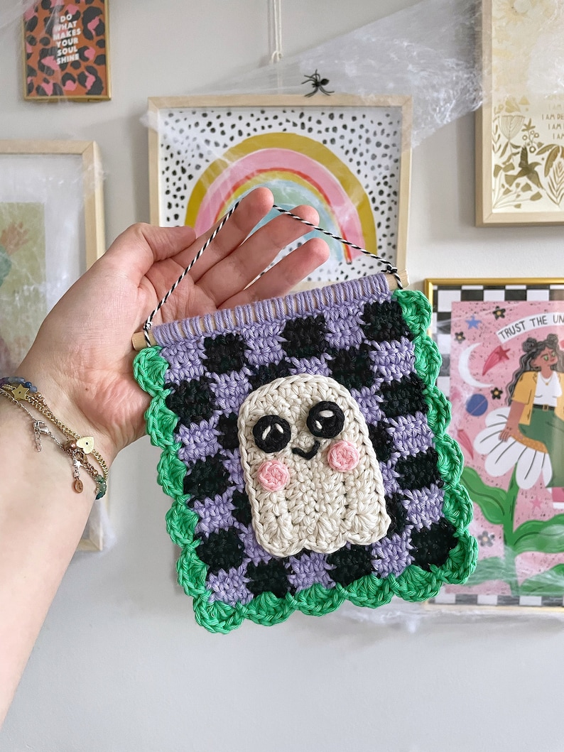 May include: Crocheted wall hanging featuring a friendly ghost design. The square banner has a checkerboard pattern in black and purple, with a green scalloped border. The ghost has black eyes and pink cheeks. A wooden dowel and string are at the top for hanging.