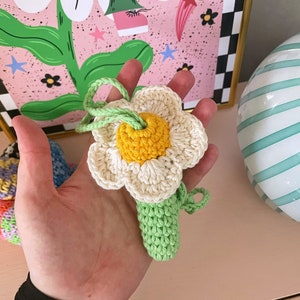 Op de afbeelding: Een gehaakte bloem met een gele kern en witte bloemblaadjes bevestigd aan een groene stengel. De bloem is handgemaakt en zou een geweldig cadeau zijn.
