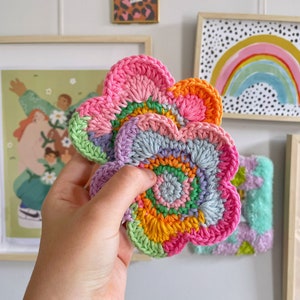 May include: Two crocheted flower-shaped coasters in shades of pink, purple, green, orange, and yellow. The coasters have a circular pattern in the centre.