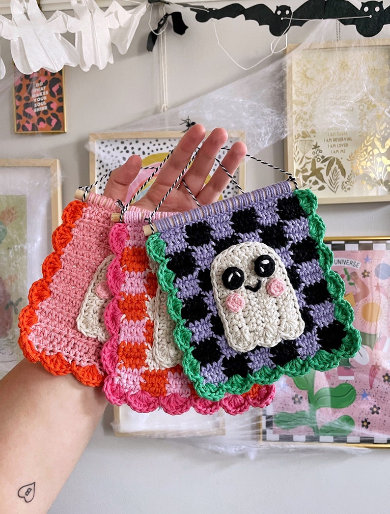 May include: A collection of handmade crochet banners in various colors, including pink, orange, and purple. One banner features a smiling ghost design. Each banner has a scalloped edge and is suspended by a black and white string.