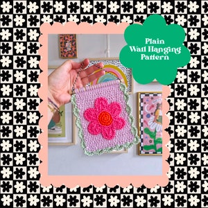 May include: A crocheted wall hanging with a pink flower in the center. The flower has a green border and a light purple background. The hanging is attached to a string and is being held by a hand.