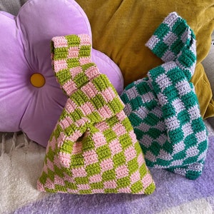 May include: Two handmade crochet bags with a checkered pattern. One bag is green and pink, the other is green and white. The bags are sitting on a purple blanket and are next to a purple flower-shaped pillow and a yellow pillow.