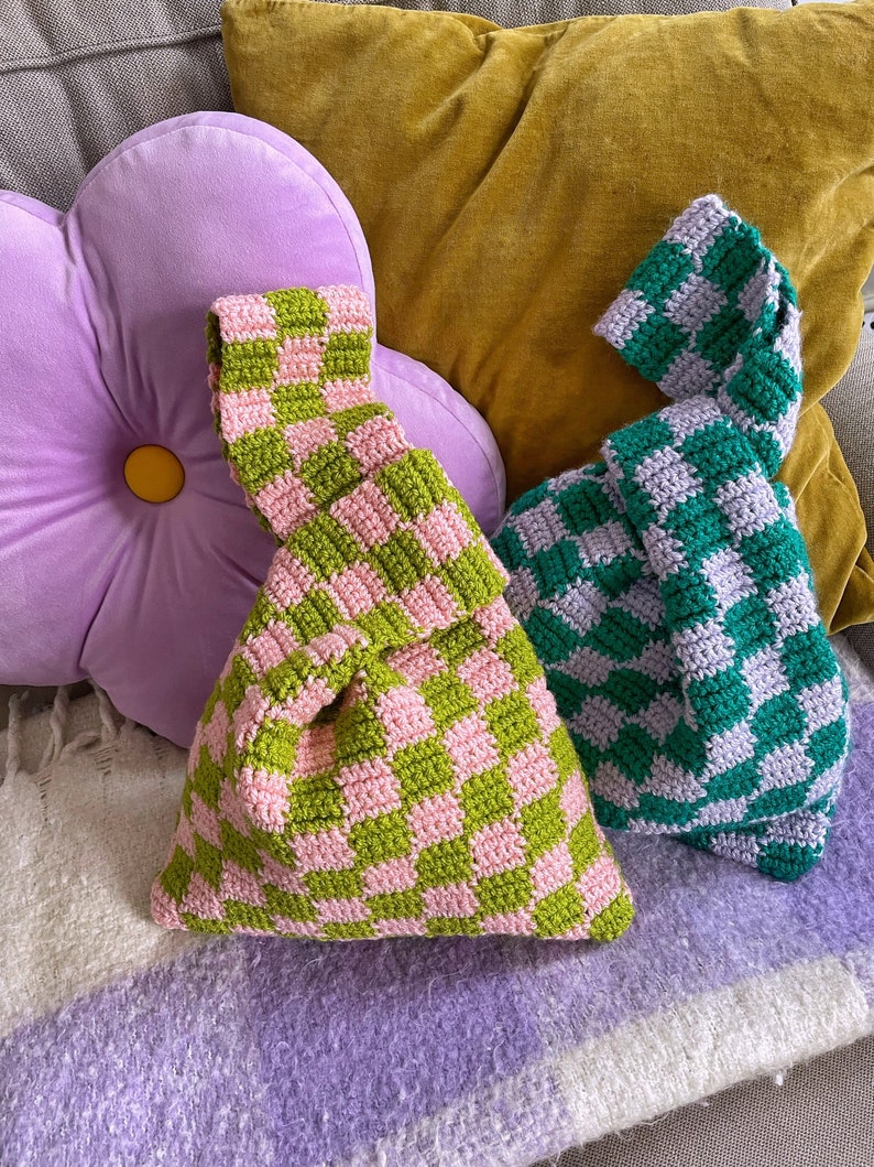 May include: Two handmade crochet bags with a checkered pattern. One bag is green and pink, the other is green and white. The bags are sitting on a purple blanket and are next to a purple flower-shaped pillow and a yellow pillow.