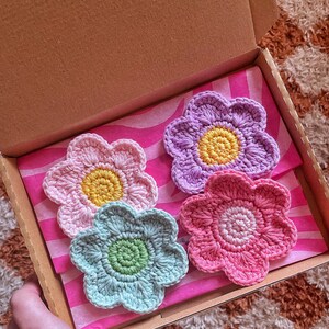 May include: A set of four handmade crochet flower coasters in a cardboard box. The coasters are in shades of pink, purple, and blue, with yellow and green centers. The box is lined with pink and white striped tissue paper.