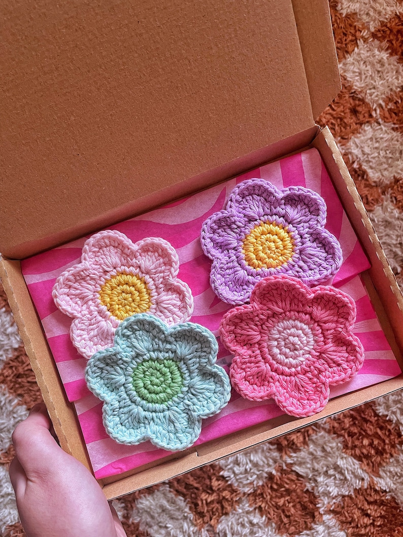 May include: A set of four handmade crochet flower coasters in a cardboard box. The coasters are in shades of pink, purple, and blue, with yellow and green centers. The box is lined with pink and white striped tissue paper.