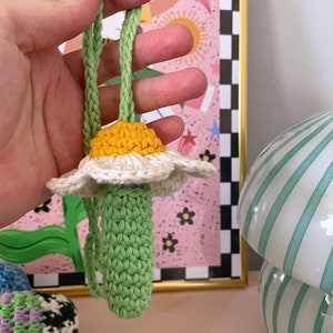 Op de afbeelding: Een gehaakte madeliefje met een groene stengel en een geel hart. De bloem is bevestigd aan een groen koord.