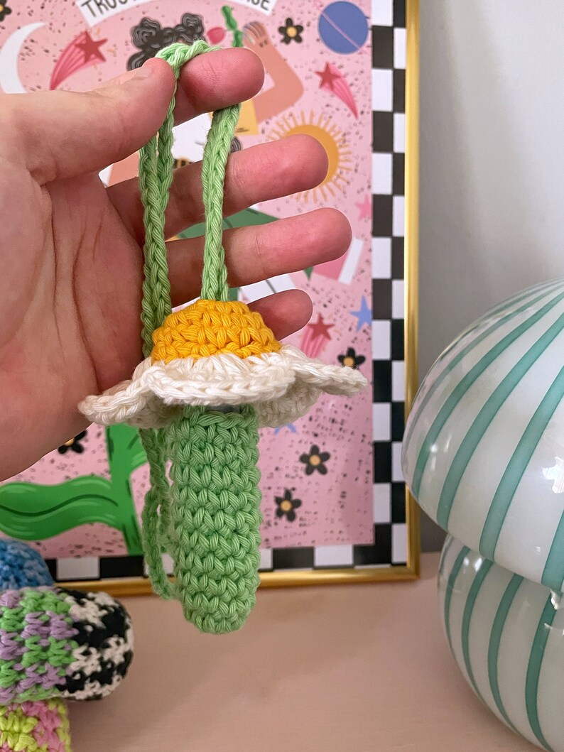 Op de afbeelding: Een gehaakte madeliefje met een groene stengel en een geel hart. De bloem is bevestigd aan een groen koord.