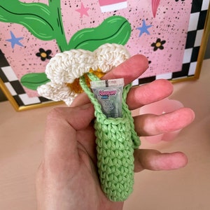 Op de afbeelding: Een groene gehaakte tas met een witte gehaakte bloem erop. De tas bevat een klein doorzichtig plastic buisje met de tekst "Powerpuff Girls" erop gedrukt.