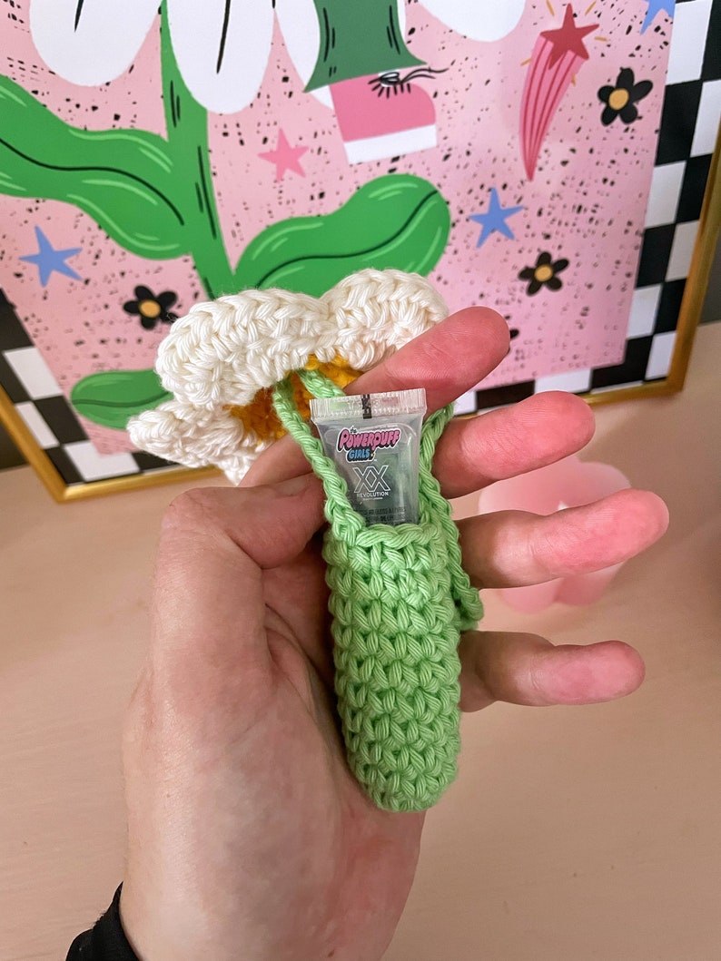 Op de afbeelding: Een groene gehaakte tas met een witte gehaakte bloem erop. De tas bevat een klein doorzichtig plastic buisje met de tekst "Powerpuff Girls" erop gedrukt.