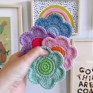 May include: A hand holds a stack of five crocheted flower-shaped coasters. The coasters are in shades of blue, purple, pink, and green, each with a contrasting center. The background includes framed art with a rainbow and text.