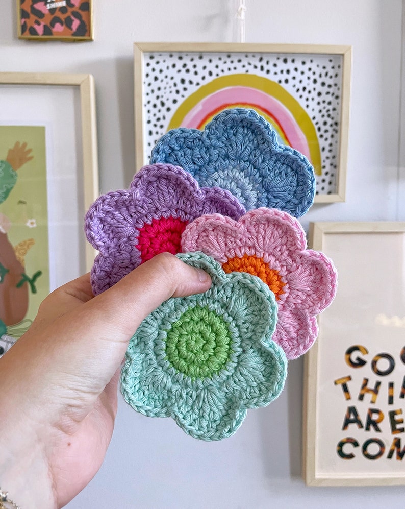 May include: A hand holds a stack of five crocheted flower-shaped coasters. The coasters are in shades of blue, purple, pink, and green, each with a contrasting center. The background includes framed art with a rainbow and text.
