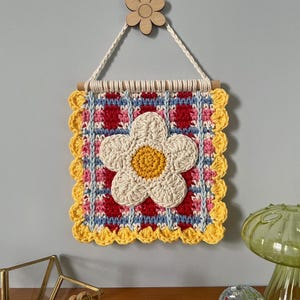 K&ouml;nnte beinhalten: Quadratischer, handgefertigter Wandbehang aus H&auml;kelarbeit mit einer gro&szlig;en wei&szlig;en G&auml;nseblume mit gelbem Zentrum. Der Hintergrund hat ein rotes, blaues und rosa Muster mit einem gelben, gewellten Rand. Oben ist eine kleine Holzblume befestigt.