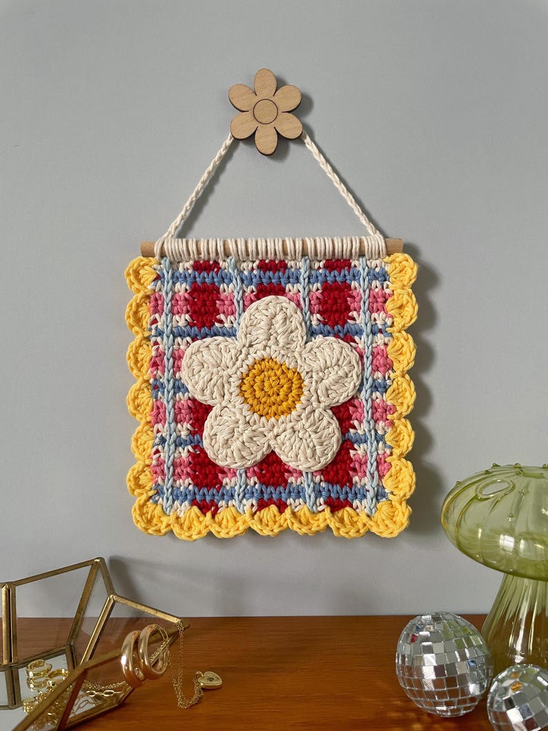 K&ouml;nnte beinhalten: Quadratischer, handgefertigter Wandbehang aus H&auml;kelarbeit mit einer gro&szlig;en wei&szlig;en G&auml;nseblume mit gelbem Zentrum. Der Hintergrund hat ein rotes, blaues und rosa Muster mit einem gelben, gewellten Rand. Oben ist eine kleine Holzblume befestigt.